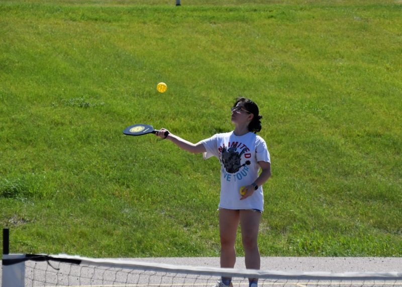 Paige plays paddle ball outdoors, hitting a yellow ball on a sunny day.