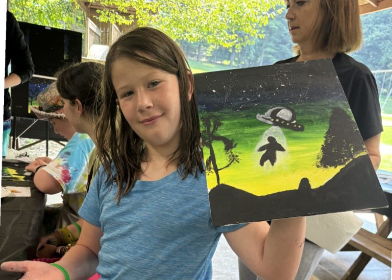 Devin smiles while holding up her painting of a nighttime scene with a UFO beaming up a figure, created during an outdoor art activity.