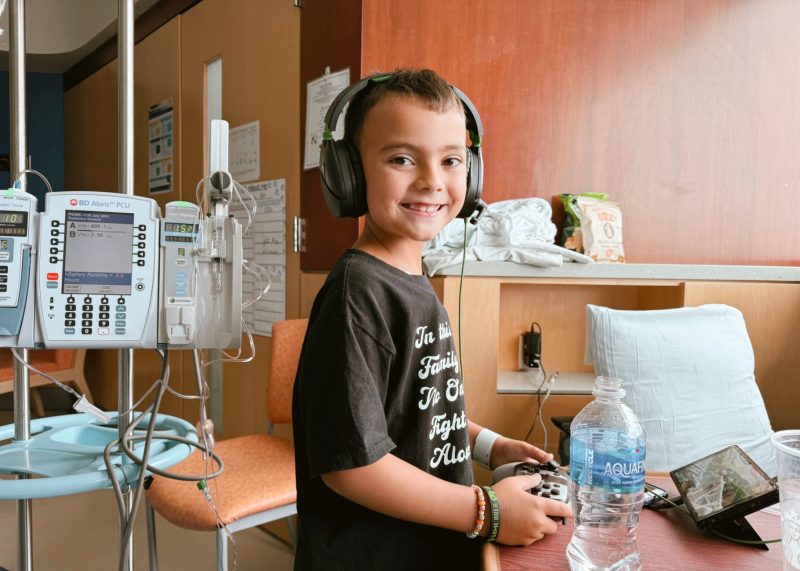 Asher is in a hospital room wearing headphones and holding a gaming controller. He is sitting next to a medical IV stand with infusion pumps, and there is an Aquafina water bottle on the table.