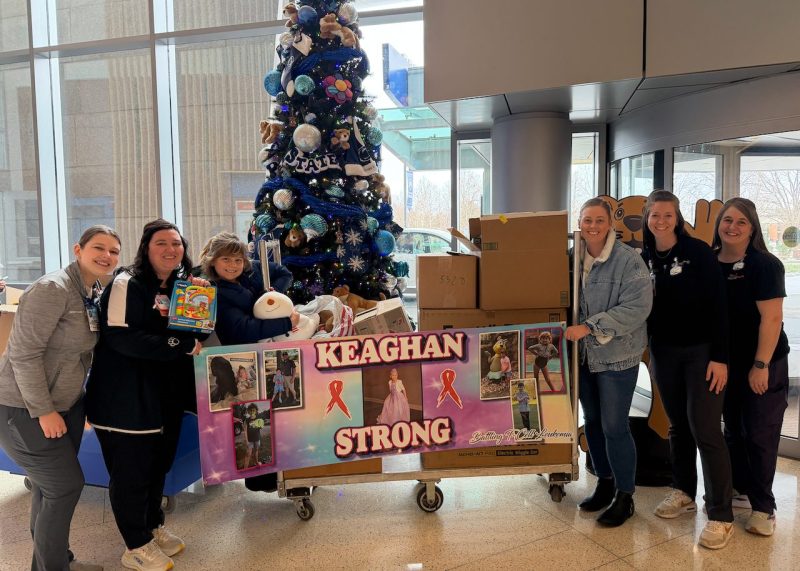 Group of people standing in a hospital lobby next to a decorated Christmas tree. They are holding a banner with 'Keaghan Strong' written on it, surrounded by boxes and toys.