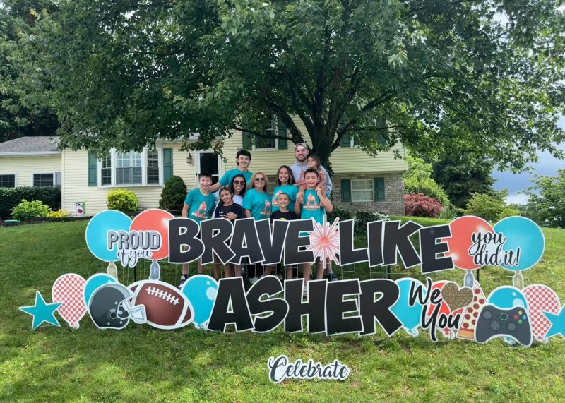 Asher and his family is standing on a lawn in front of a large sign that reads, "Proud of you, Brave like Asher, You did it, We love you, Celebrate." The sign includes images of balloons, a football, and a game controller. There is a large tree and a house in the background.