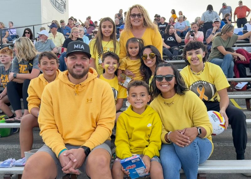 Asher and his family are sitting on stadium bleachers and wearing yellow and black-themed clothing. Some hold sports-themed items and have face paint. A large crowd is visible in the background. The atmosphere suggests a sports event.