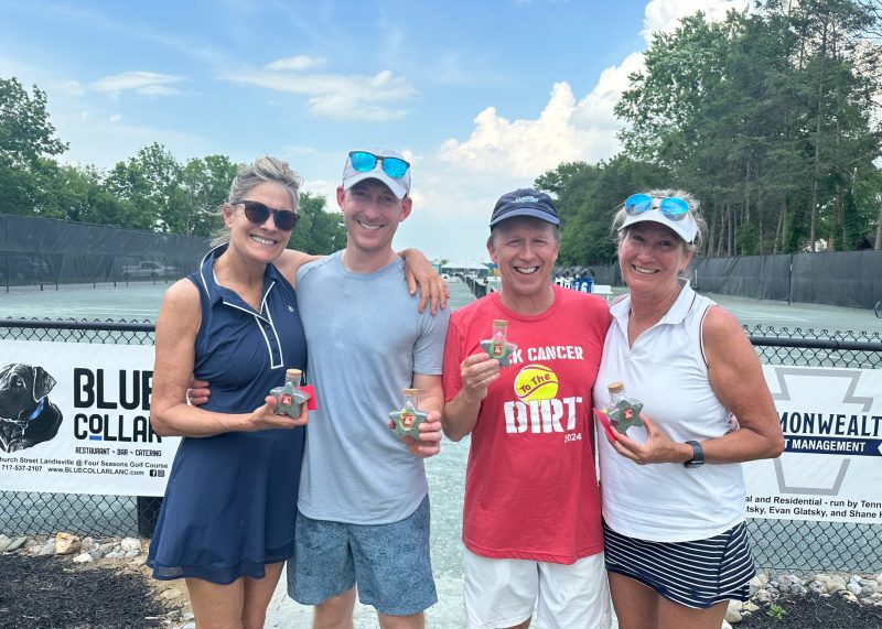 Four people smiling and holding small trophies stand in front of a tennis court. They appear to be enjoying a sunny day outdoors.