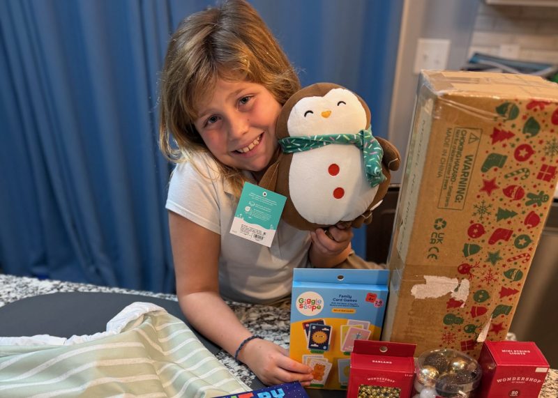 A person smiling while holding a penguin plush toy. Displayed on the surface are a cardboard shipping box, a "Paint by Sticker Kids Christmas" book, "The Night Before Christmas" book, and several holiday-themed packages and decorations.
