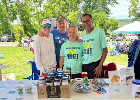 A group of four people is standing behind a table covered with various items and literature. The table has a prominently displayed Four Diamonds banner with the slogan "Conquering Childhood Cancer." The setting is outdoors with trees and a grass field in the background.
