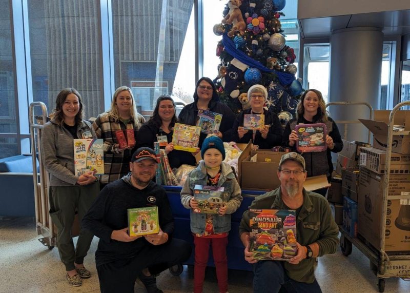 A group of individuals is gathered in a hospital lobby, holding various toys, with a decorated Christmas tree in the background. Numerous boxes are visible, likely containing more toys, and there is a teddy bear decoration nearby.