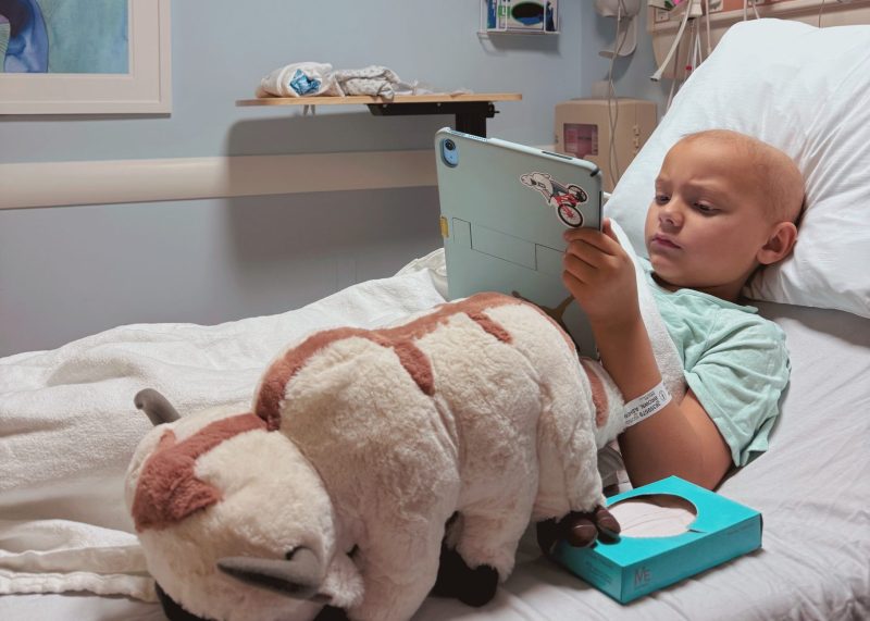 Asher is in a hospital bed engaged with a tablet. Next to him is a large stuffed animal resembling Appa from "Avatar: The Last Airbender". The room has medical equipment and a framed picture on the wall.
