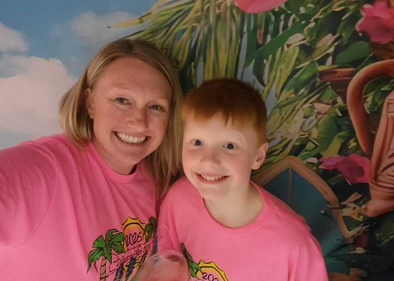 Sean and his mom, Melissa, smile together in matching pink shirts at a Mini-THON® event, posing in front of a colorful tropical backdrop.