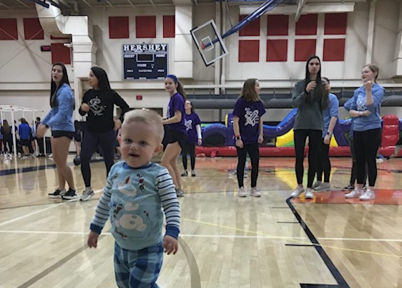 Daniel walks across a gym floor in the foreground while students dance and participate in a Mini-THON® event behind him, with a “HERSHEY” scoreboard and inflatable activities visible in the background.