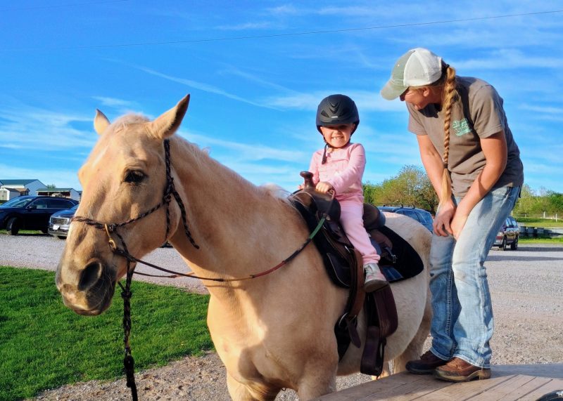 A person guides Bella sitting on a light brown horse. Bella is wearing a helmet and is smiling. There are parked cars and a clear blue sky in the background.