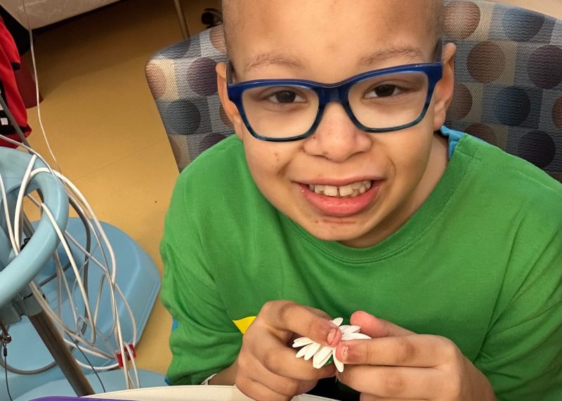 Elias smiles while holding a white flower during a hospital visit. He wears blue glasses and a green shirt.