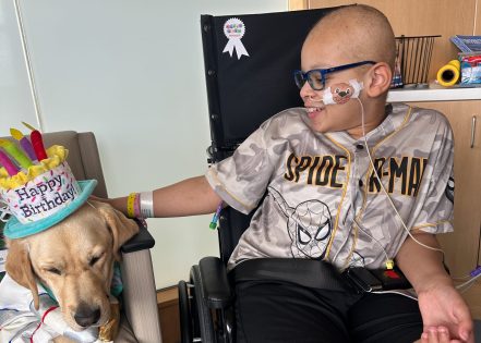Elias smiles while sitting in a wheelchair beside a therapy dog wearing a birthday hat. He gently pets the dog during a hospital visit.
