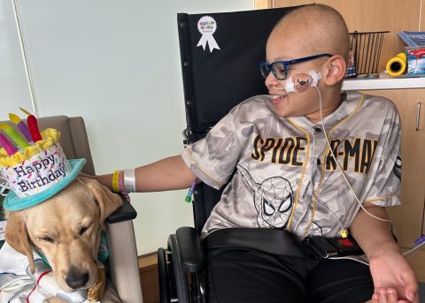 Elias smiles while sitting in a wheelchair beside a therapy dog wearing a birthday hat. He gently pets the dog during a hospital visit.