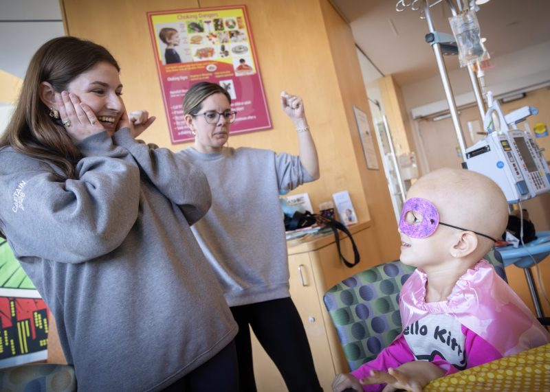Presley, a Four Diamonds child, wearing a superhero mask and cape smiles with two visitors during cancer treatment in a hospital room.