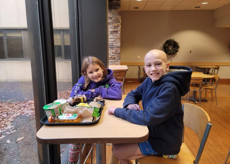 Asher, an 11-year-old with childhood neuroblastoma, smiles with his sister while sitting together at a table.
