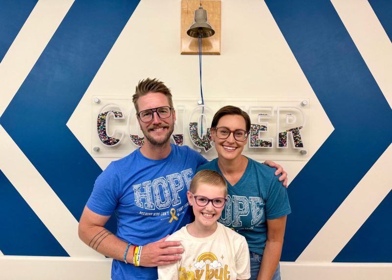 Lenny stands smiling with her parents beneath the Conquer bell following childhood cancer treatment.