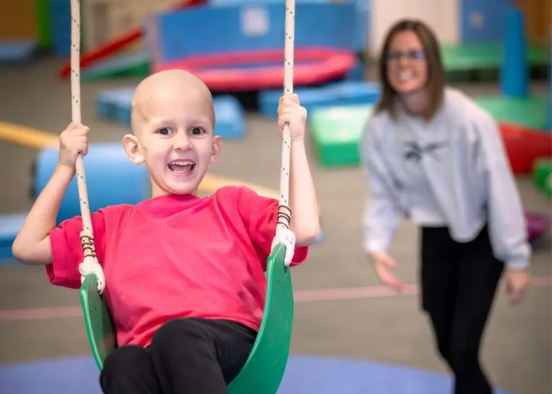 Four Diamonds child Presley, who is undergoing treatment for a Wilms tumor, smiles while playing on a swing with a staff member nearby.