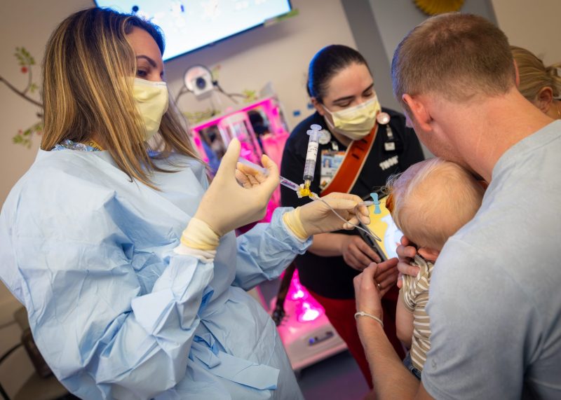 A care team administers treatment to a young pediatric cancer patient being comforted by a parent.