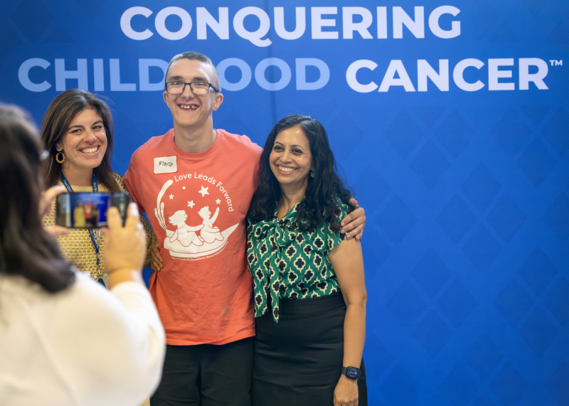 Three people smile for a photo in front of a blue Four Diamonds “Conquering Childhood Cancer” backdrop while another person takes their picture.