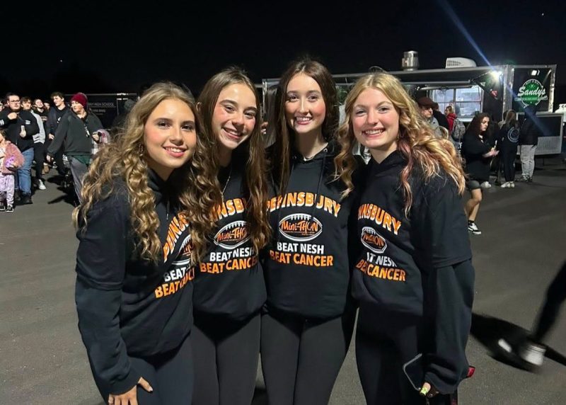 Four Pennsbury High School students smile together while wearing matching black Mini-THON hoodies that read “Beat Neshaminy, Beat Cancer” at an outdoor nighttime event.