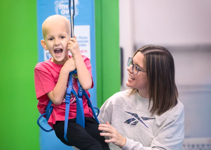 Four Diamonds child Presley smiles while using a therapeutic swing with support from a care provider during a therapy session.