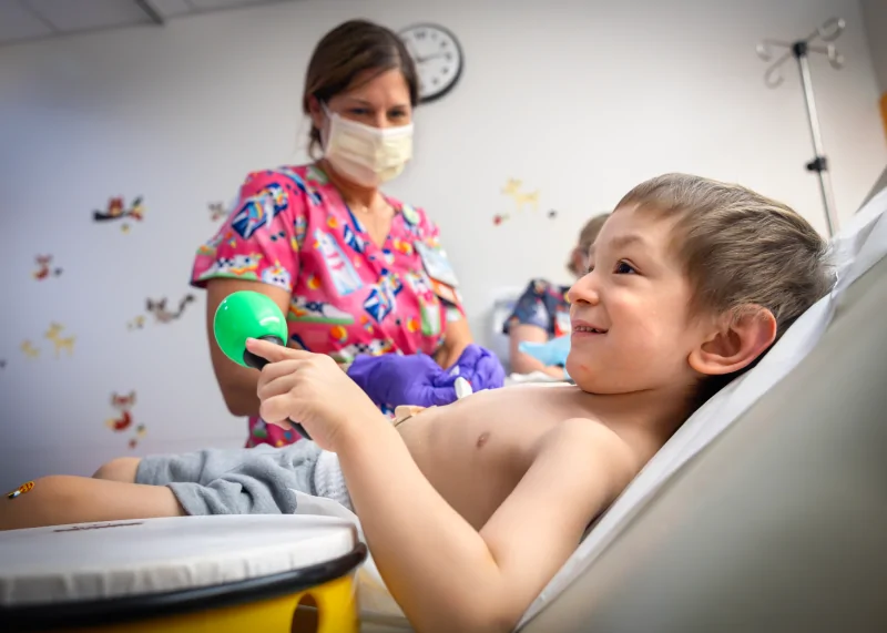 A nurse is tending to Brayden on a hospital examination bed. Brayden is relaxed and receiving care. The room is equipped with medical supplies and has playful decor on the walls.