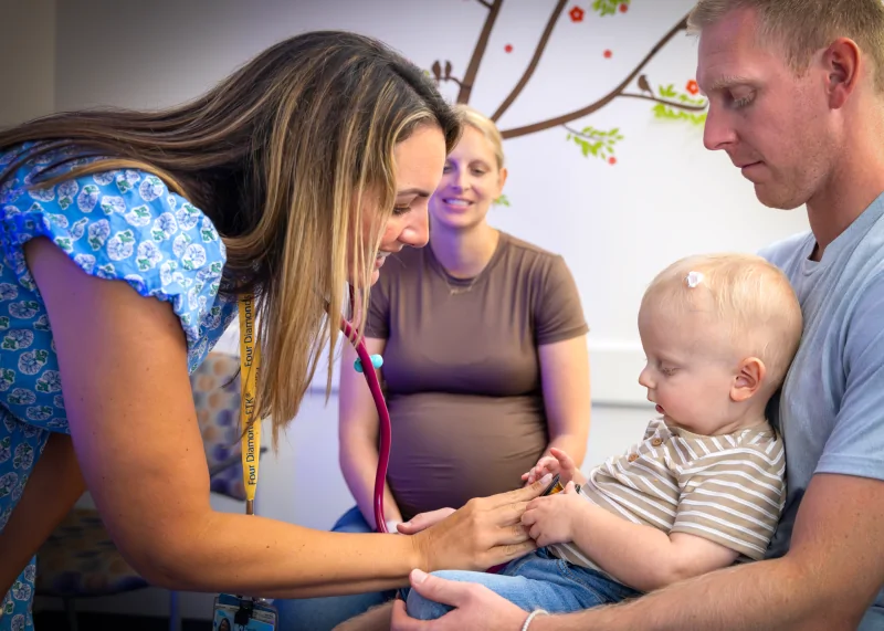 Christina, a nurse practitioner, uses a stethoscope on Harlan sitting on his dad's lap. His mom is seated nearby. The setting appears to be a medical office with colorful wall decorations.