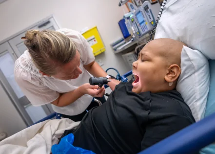 A healthcare worker examines Tim in a hospital bed, using a medical instrument to check his mouth. Medical equipment is visible in the background. The setting appears to be a hospital room.