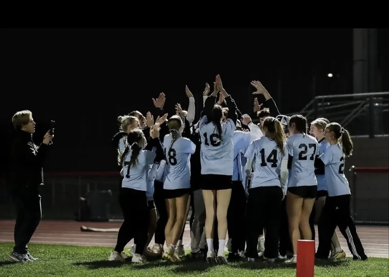 Annville High School students wearing white jerseys gather in a team huddle with their hands raised during a nighttime powder puff football game.